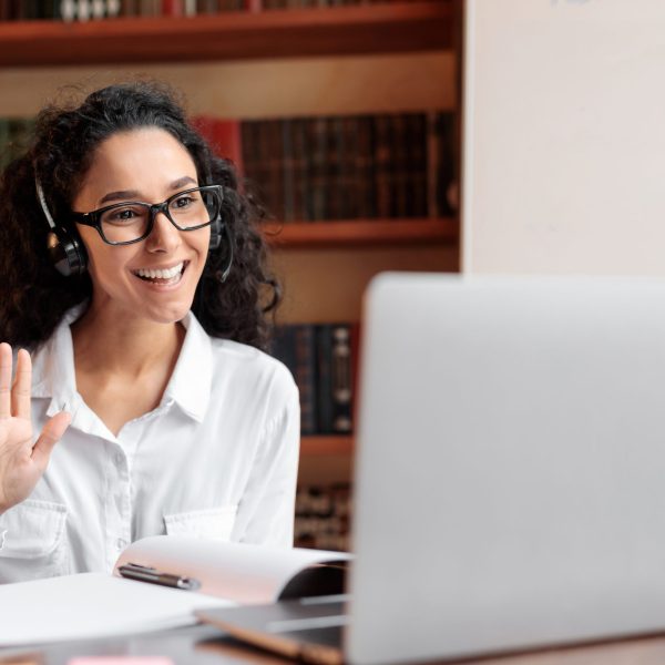 Online Tutoring. Portrait Of Woman In Glasses And Headset Having Video Conference, Sitting At Desk At Hone Office, Learning Foreign Languages, Waving To Camera During Web Call, Greeting Indian Teacher